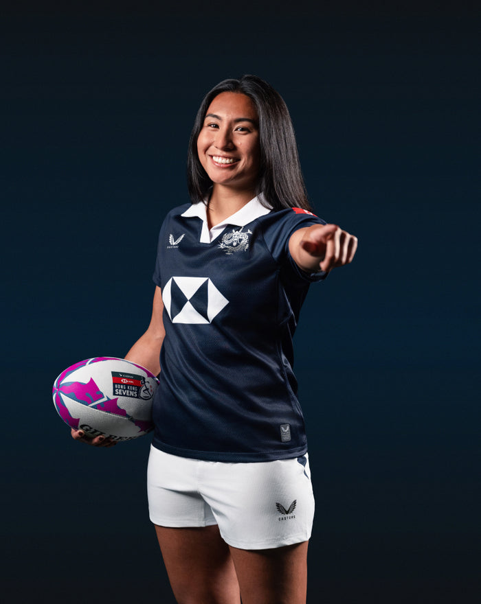 Female model wearing navy Hong Kong Sevens home replica jersey holding a rugby ball and smiling.