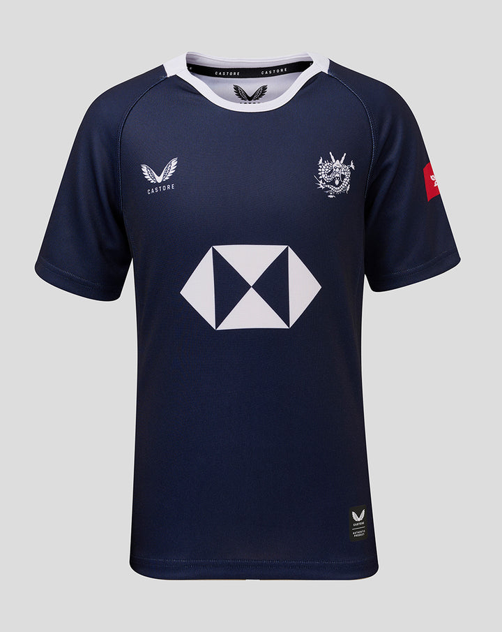 Front view of navy junior home replica jersey with white collar, team crest on chest, Castore branding, and a large geometric sponsor logo in the center.